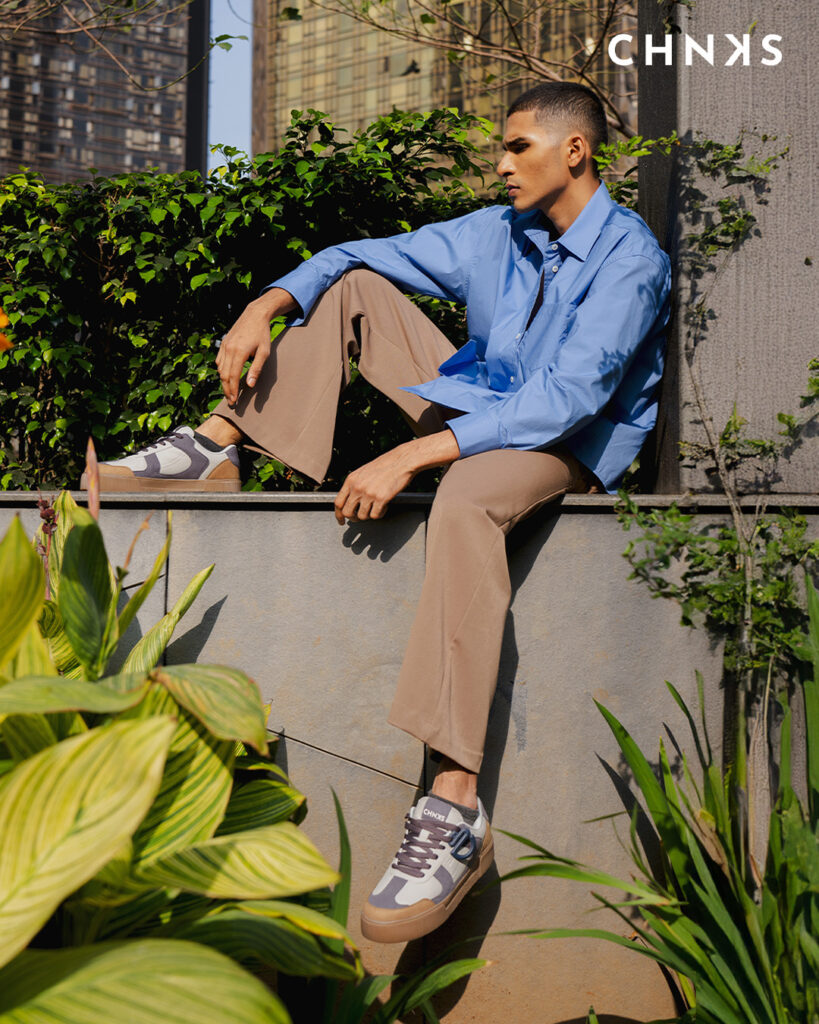 Man seated on a garden terrace in blue shirt and beige trousers wearing CHNKS low-top men sneaker in granite grey and cream with gum sole; clean minimal panels, padded tongue, detachable charm, modern city towers behind lush greenery.