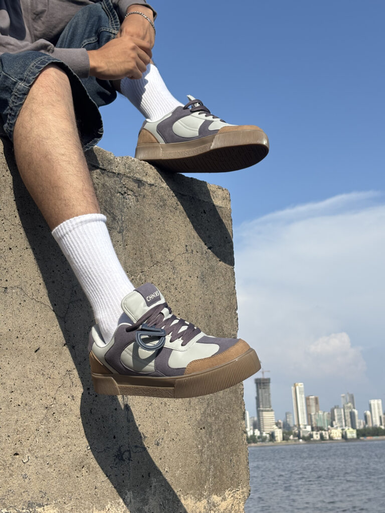 Man on a concrete ledge by the waterfront skyline wearing CHNKS low-top men sneaker in granite grey, cream and gum brown; detachable CHNKSHEAD token charm, padded tongue, vulcanized gum sole, casual streetwear shot in natural light