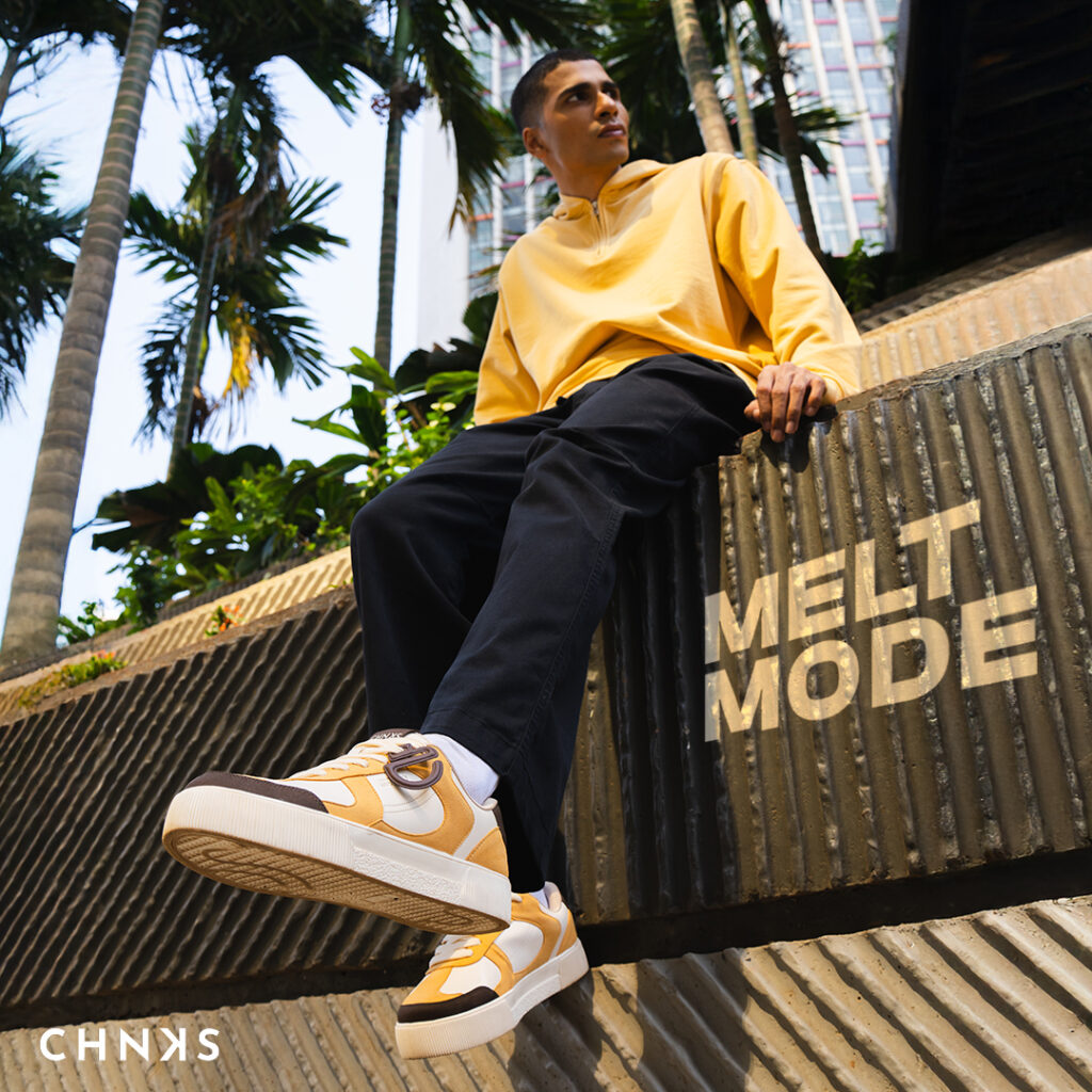 Urban lifestyle shot of a man in a yellow sweatshirt wearing CHNKS low-top men sneaker in cream and mustard with gum sole; detachable CHNKSHEAD token charm visible, outsole tread detail, palm trees and city architecture in the background.